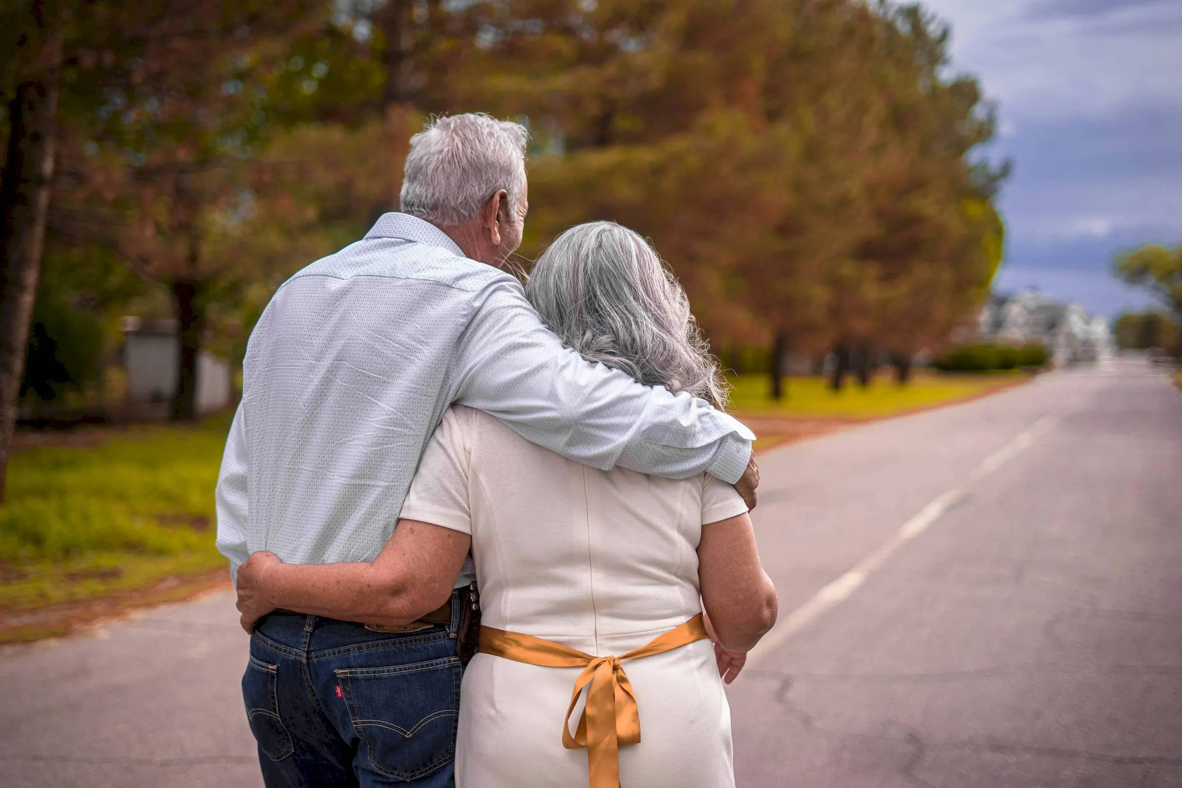 An elderly couple walks arm in arm down a tree-lined road, honoring every story with each step they take together.
