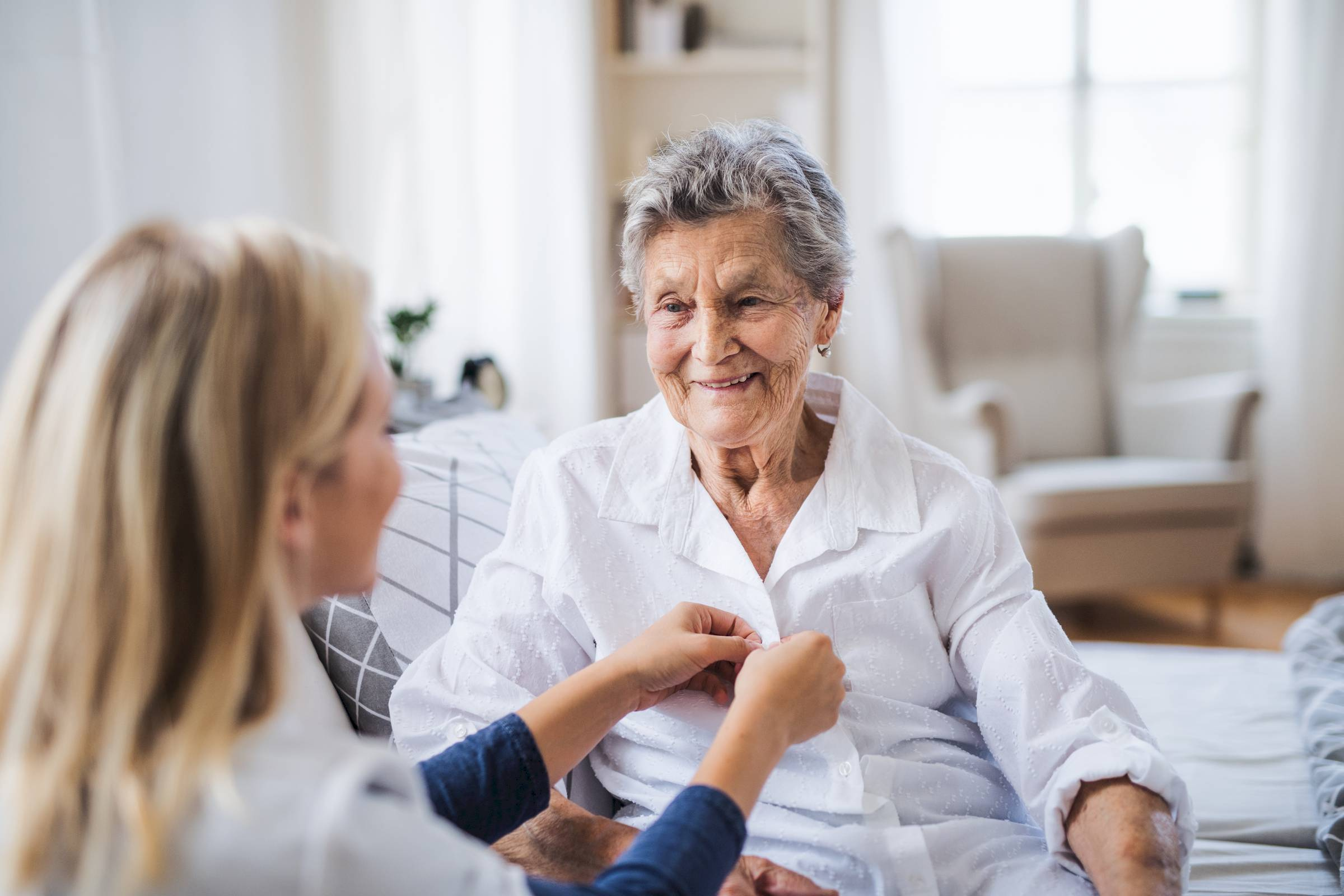 Young woman helps an elderly woman button her shirt as they sit together on a couch in a bright living room.