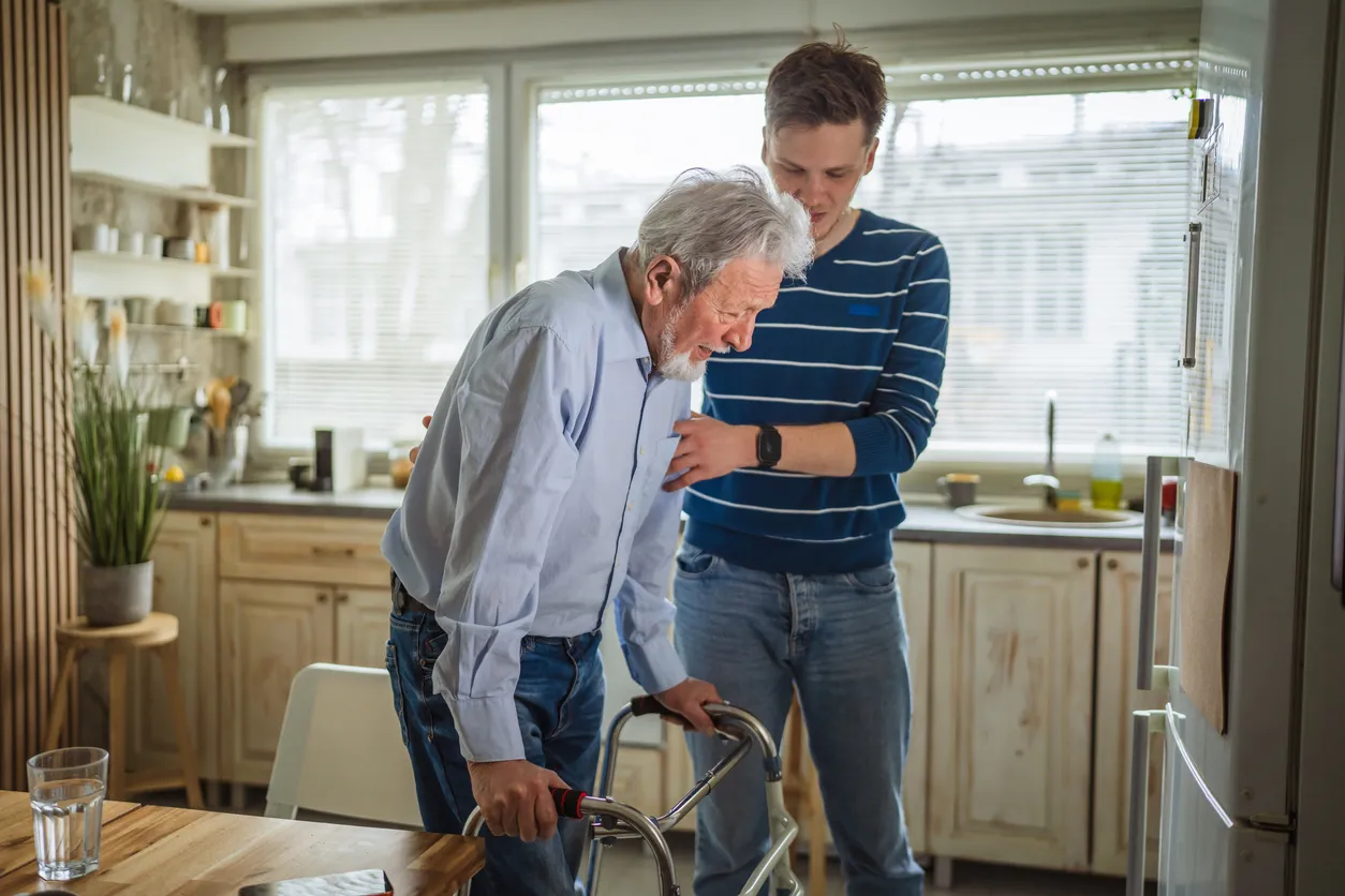 Young man helps elderly man with a walker in a bright kitchen, discussing signs of assisted living and future care options.